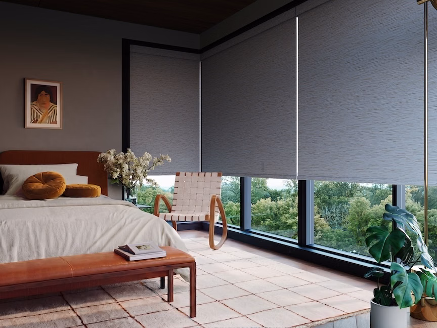 Dark blackout roller shades cover floor-to-ceiling bedroom windows above a wood bench and neutral area rug.