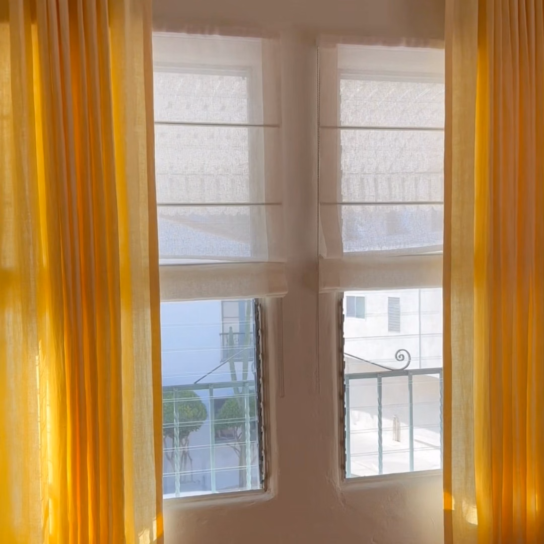 White sheer linen relaxed Roman shades layered with Buttercup drapes in a bright living room.